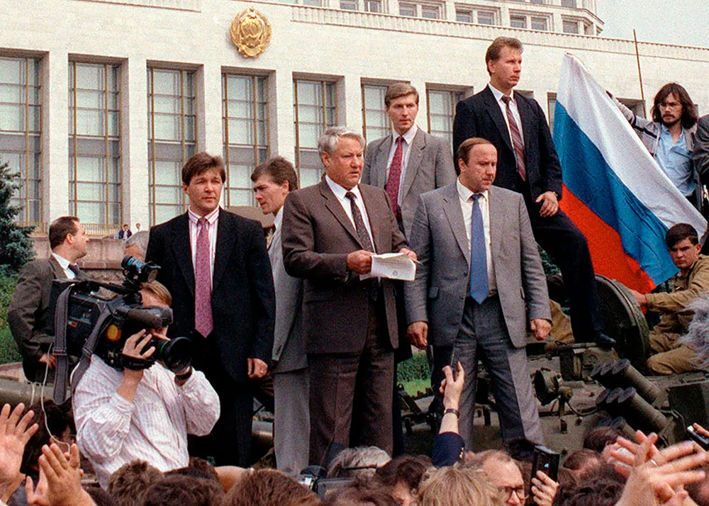 Russian men in suits and ties stand on a tank while a crowd cheers around them. In the background is the Russian Parliament building, a huge white building with many windows, some open.