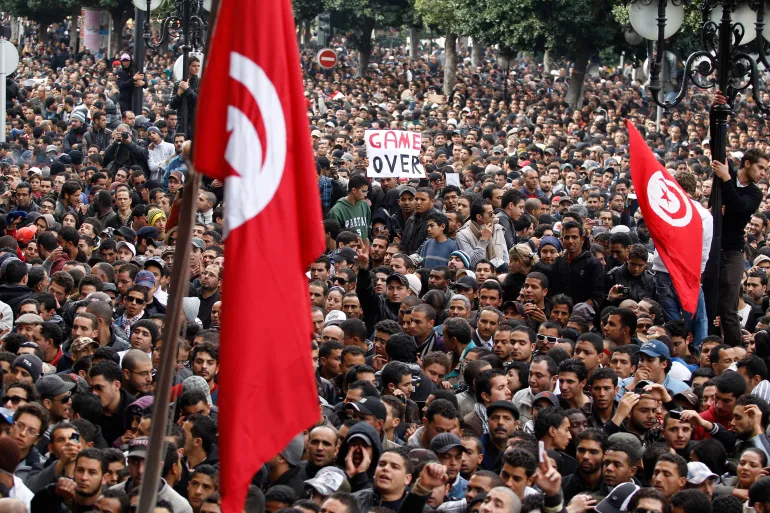 Crowd of protesters with the Tunisian flag in the foreground. There is a sign that reads "Game Over".