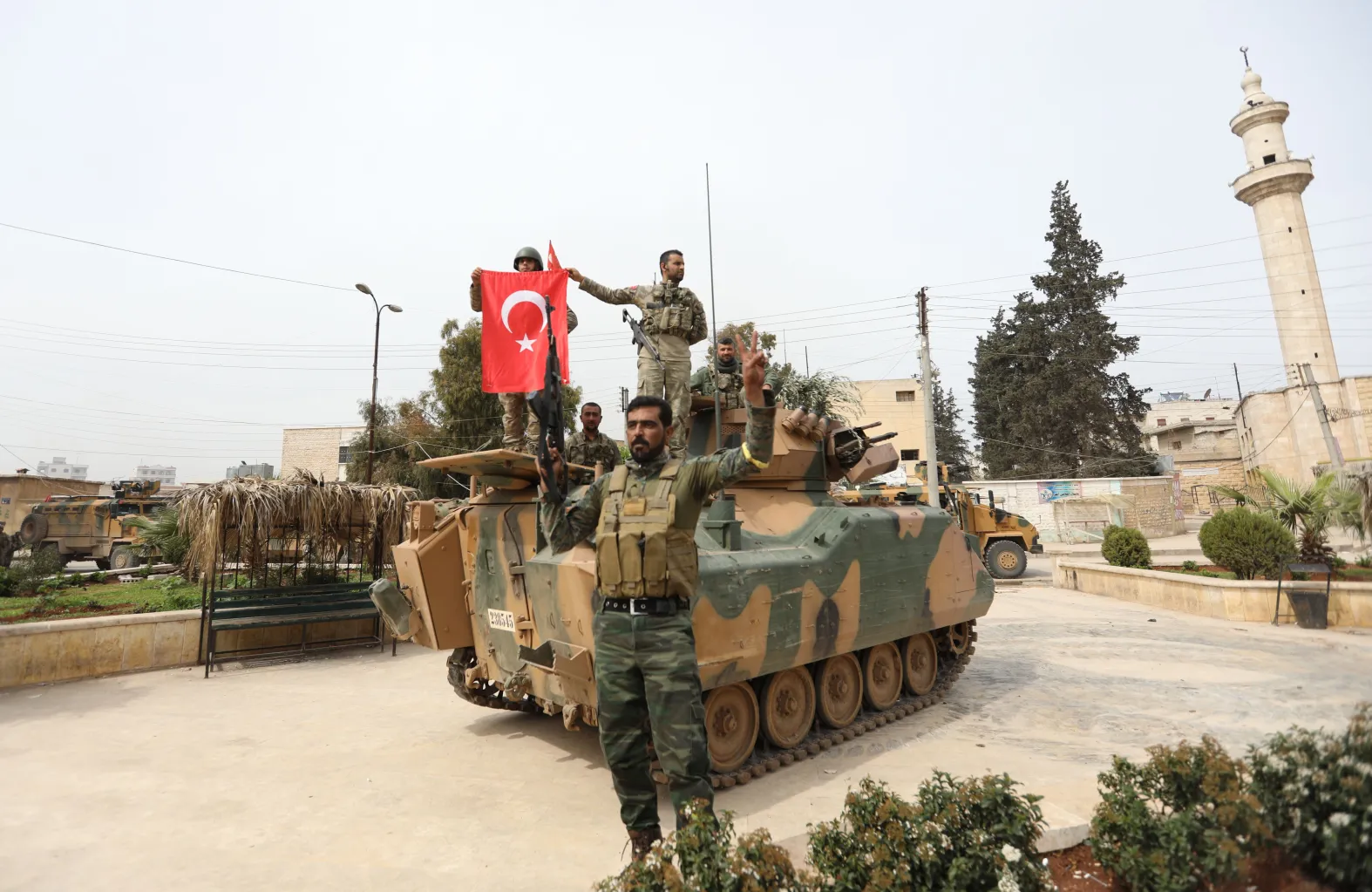 Men with dark hair and beards in green camouflage uniforms stand and sit on a camouflaged tank waving a Turkish flag (white crescent and star on red background). The man standing in front of the tank points his machine gun toward the sky. There is a building with a white town on the right side of the picture.