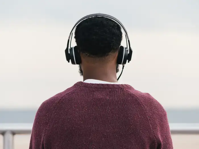 The back of a Black man with short, curly black hair, a curly black beard visible. He's wearing over-the-ear black headphones a brownish-red sweater with a white shirt peaking out at the collar.