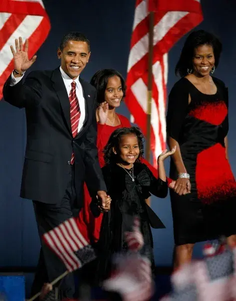 A Black family, including a man, a woman, and two girls, wave to a crowd waving American flags. He is wearing a dark suit with a red and white stripped tie; the woman is wearing a black and red dress; the taller girl is wearing a red dress and holds the woman's hand, and the shorter girl is wearing a black dress and holding the man's hand. The man and both girls are waving at the crowd. All four are smiling broadly.