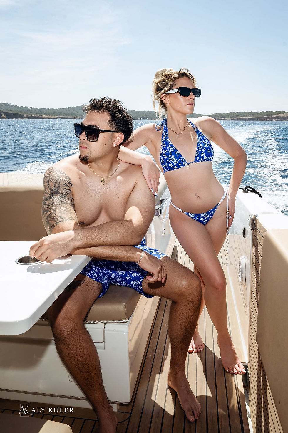 Couple in blue swimwear and sunglasses posing on a boat with ocean and shoreline behind them;