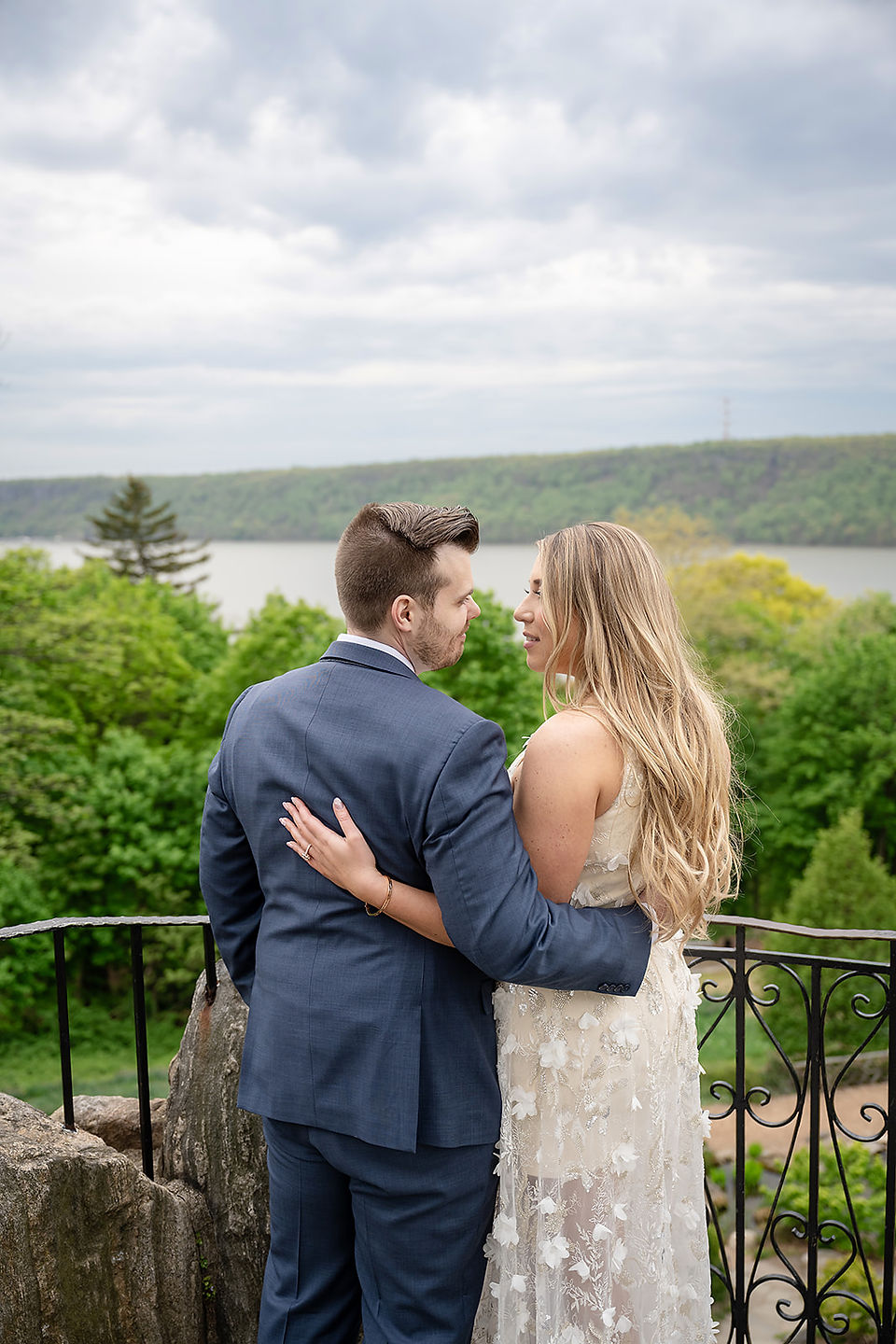 A couple embraces by a pond in a garden, the woman in a white dress and the man in a blue suit. Greenery and cloudy sky in the background.