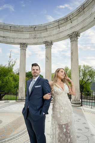 A couple embraces by a pond in a garden, the woman in a white dress and the man in a blue suit. Greenery and cloudy sky in the background.