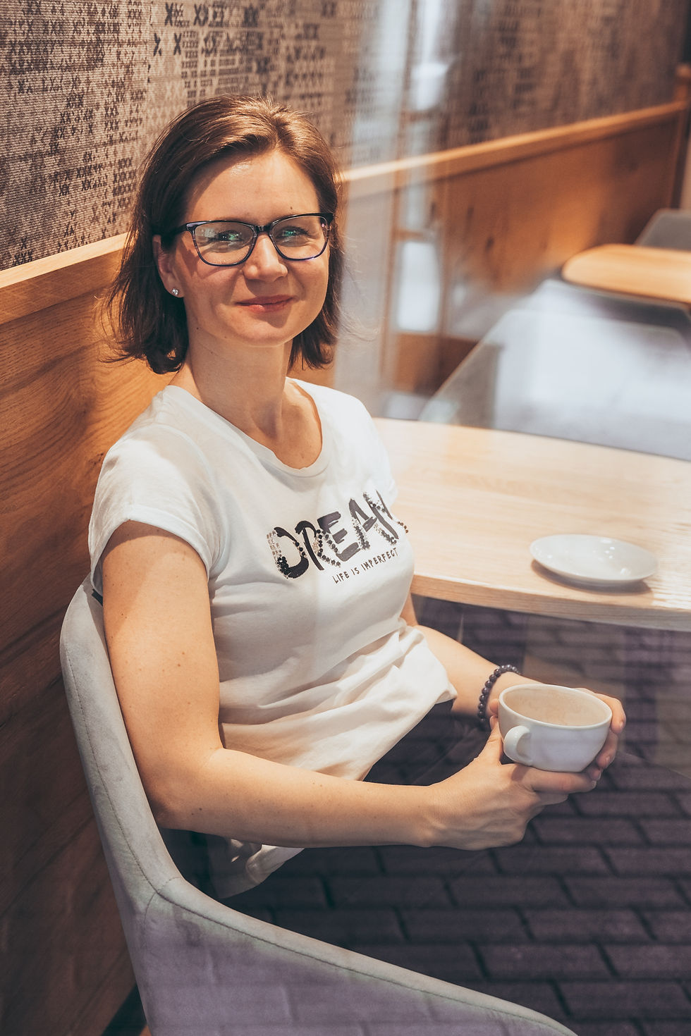 Miroslava smiling gently over coffee in a relaxed café setting, wearing a shirt that reads “DREAM — Life Is Imperfect,” embodying realness and everyday leadership.