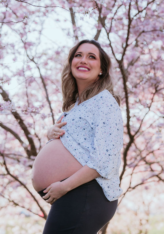 Stunning mama-to-be shines under the soft pink cherry blossoms, her beautiful smile lighting up the scene as she gently holds her big bump, which peeks out from beneath her shirt, embracing the beauty of this special moment.