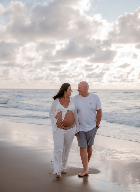 "A peaceful sunset stroll by the water in Kijkduin – mom and dad-to-be walking barefoot, dressed in light white clothes, embracing the calm beauty of the moment as they prepare for the new chapter of their lives.