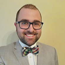 A caucasian man with short cropped brown hair and beard wearing glasses, a multi-colored plaid bow tie, and a grey suit posing in front of a tan wall.