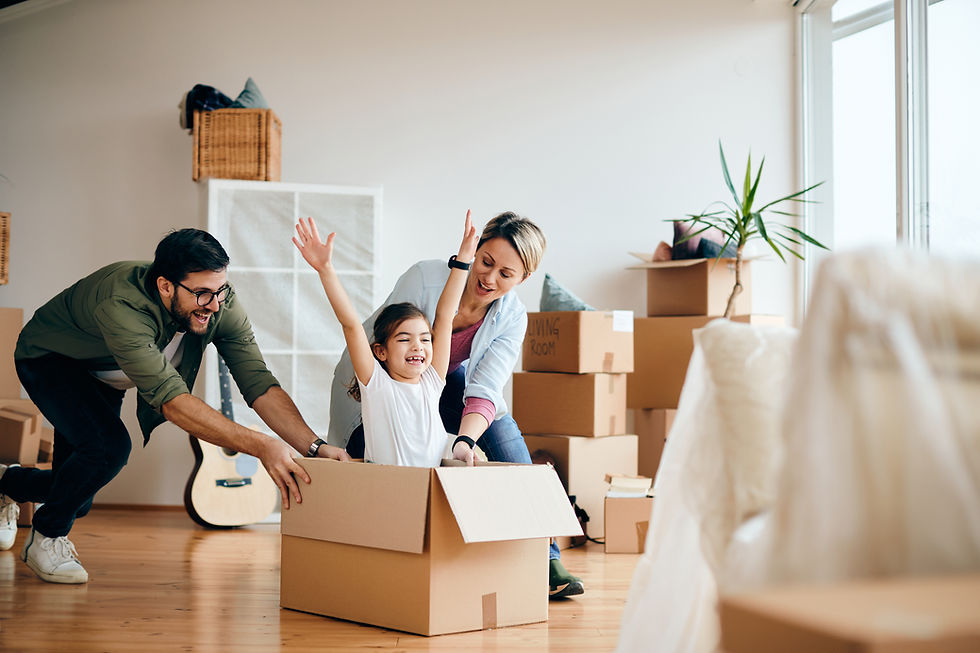 Familia sonriente jugando en una caja de mudanza en una sala luminosa, con cajas apiladas, planta y ventana al fondo.