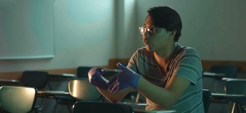 Man in glasses and blue gloves gesturing in a classroom