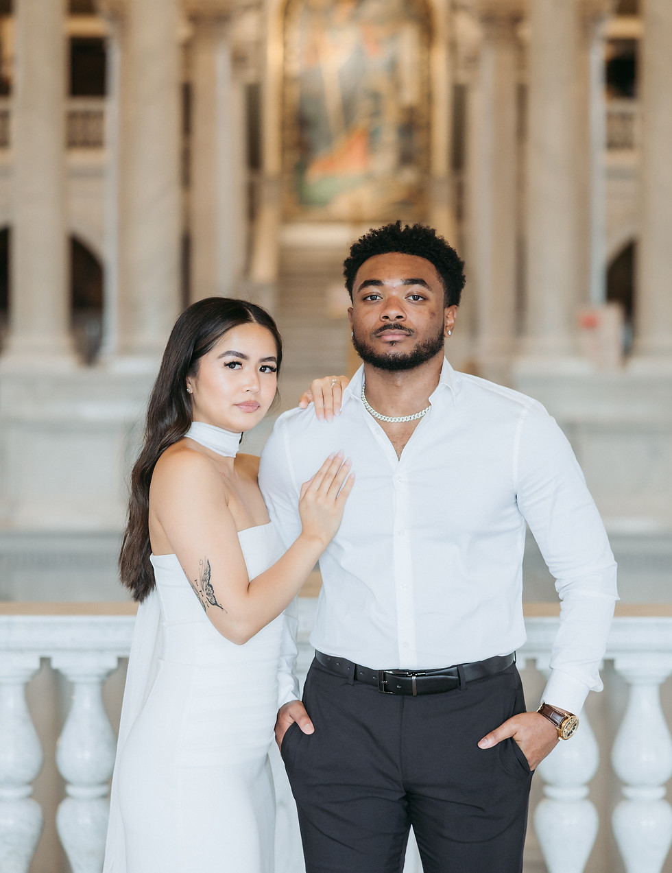 Woman in white dress and man in white shirt pose in a grand hall.