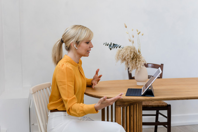 Blonde woman with a pony tail and ear pods sitting on a desk and talking to someone on the mac book online