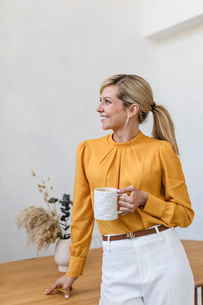 Blonde woman with a pony tail, white trousers and dark yellow top holding a coffee mug, looking to the side and smiling
