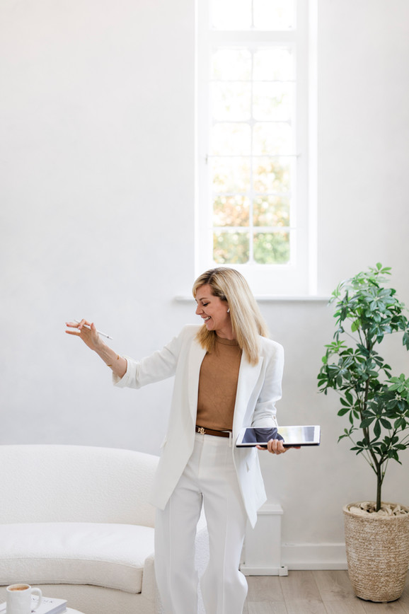 Blonde woman in a white suit holding an ipad, smiling and dancing in a bright room with a white sofa behind her