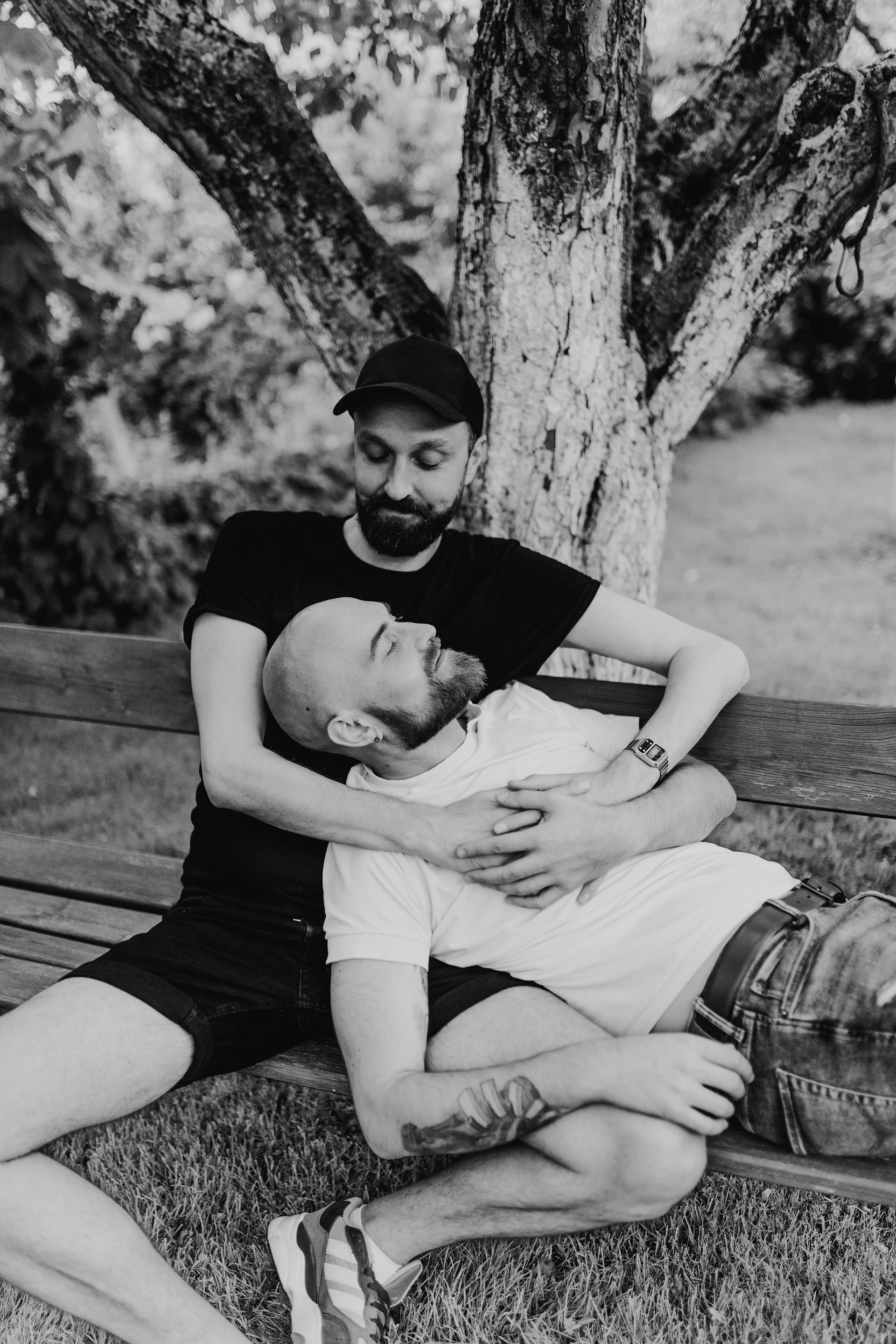 black and white photo of a gay couple , one is leaning on the other with eyes closed, sitting on a bench in front of a tree