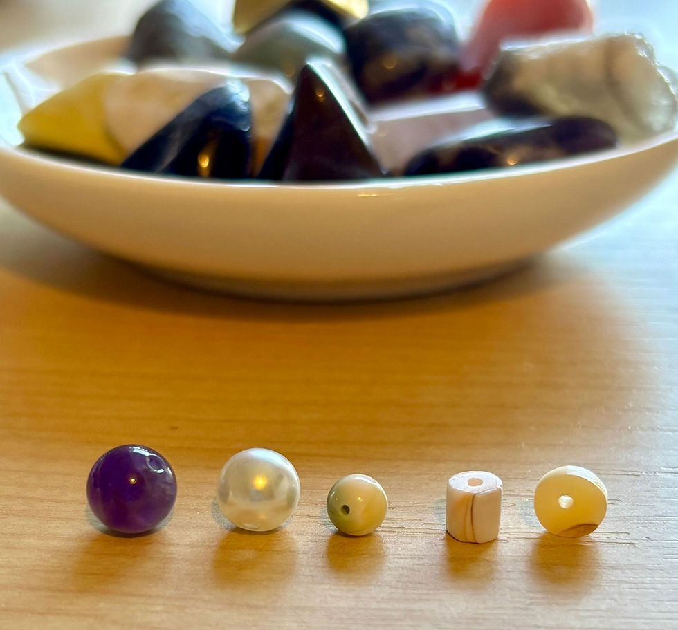 Five small beads, various colors and shapes, are lined up on a wooden surface. A bowl with stones is blurred in the background.