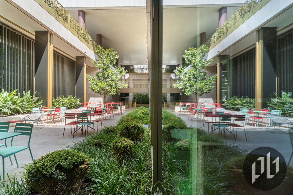 Pátio interior envidraçado com jardim central, arbustos e mesas coloridas para refeições ao ar livre.