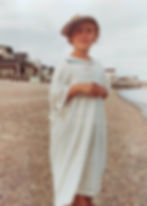 Child on a beach wearing an oversized white shirt and a sideways cap. Houses and calm sea in the background. Overcast sky. Relaxed mood.