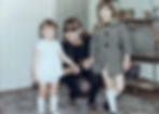 Woman kneeling between two smiling children indoors; one child in white, the other in plaid coat. Vintage TV and books in background.
