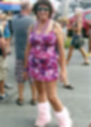 Woman in pink camo dress and furry boots smiles at outdoor event. Crowd and colorful umbrella in background, festive mood apparent.