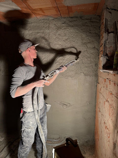 Handwerker beim Verputzen einer Innenwand: Er trägt mit einer Kelle frischen Mörtel auf eine unverkleidete Ziegelwand auf und hält ein Kellebrett in der anderen Hand. Baustellensituation im Innenraum mit sichtbaren Ziegelsteinen.