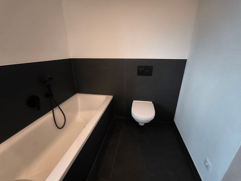 Eine weiße Badewanne und eine weiße Toilette. im Hintergrund ist alles schwarz gefließt.