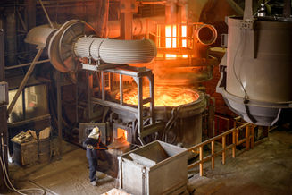 Worker in protective gear handles a furnace in an industrial manufacturing setting with glowing orange molten metal and machinery around.