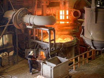 Worker in protective gear handles a furnace in an industrial manufacturing setting with glowing orange molten metal and machinery around.