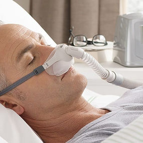 Homem dormindo com uma máscara nasal conectada a um aparelho CPAP, equipamento usado no tratamento da SAOS — Síndrome da Apneia Obstrutiva do Sono. Ele está deitado de lado na cama, em um ambiente domiciliar, com o equipamento visível ao fundo.