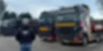 Le chef d'entreprise est de dos sur cette image en premier plan avec une doudoune sans manches floquée avec le logo et le nom de marque. En second plan ; différents camions trak trans floqués sont alignés.