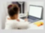A young Black girl with curly hair tied back is seated at a desk, looking at a laptop and writing in a notebook