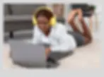 A young Black woman with braided hair and yellow headphones is lying on a light-colored rug, smiling as she looks at a laptop