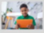 A cheerful young boy in a green shirt sitting on the floor and using a bright orange tablet in a modern, well-lit study space