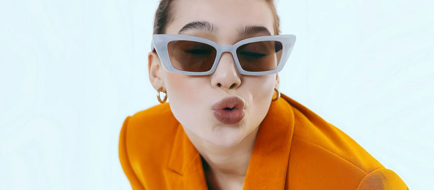 Stylish woman in white cat-eye sunglasses, orange blazer, and gold hoops, playfully blowing a kiss against a light blue background.