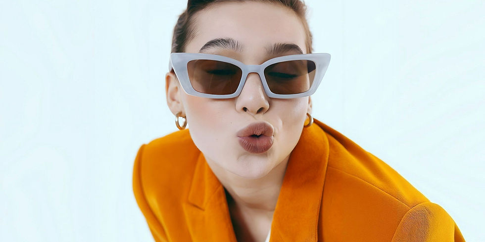 Stylish woman in white cat-eye sunglasses, orange blazer, and gold hoops, playfully blowing a kiss against a light blue background.