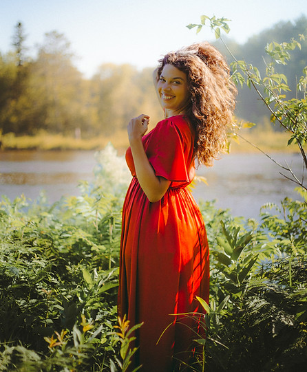 The photographer smiling and wearing a red dress while standing in tall grasses and plants.