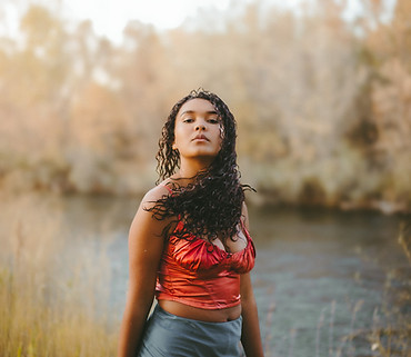 A young woman with an empowered expression. She is near a river and is whipping her wet curls to the side.