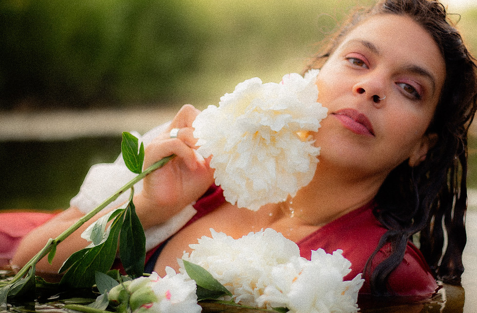 The photographer posing for an image. She is in water wearing a red dress and holding white flowers.