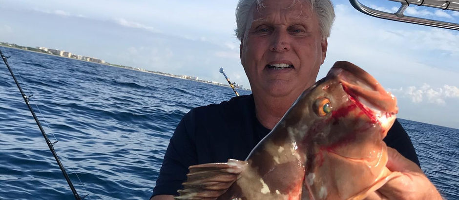 Man on boat holding a large fish with speckled scales. Blue ocean and sky in background, fishing rods visible. Appears pleased.