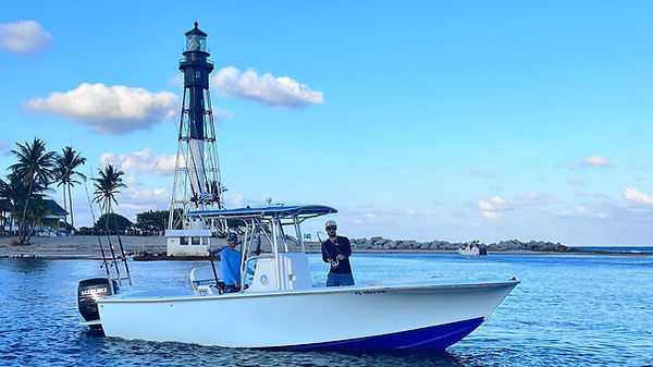 a man driving a boat in the intercoastal and another man fighting a fish with the rod bent over the bow while the fish is pulling out line in the backgroud there is another small boat on a beach with rocks in the distance and a tall light house