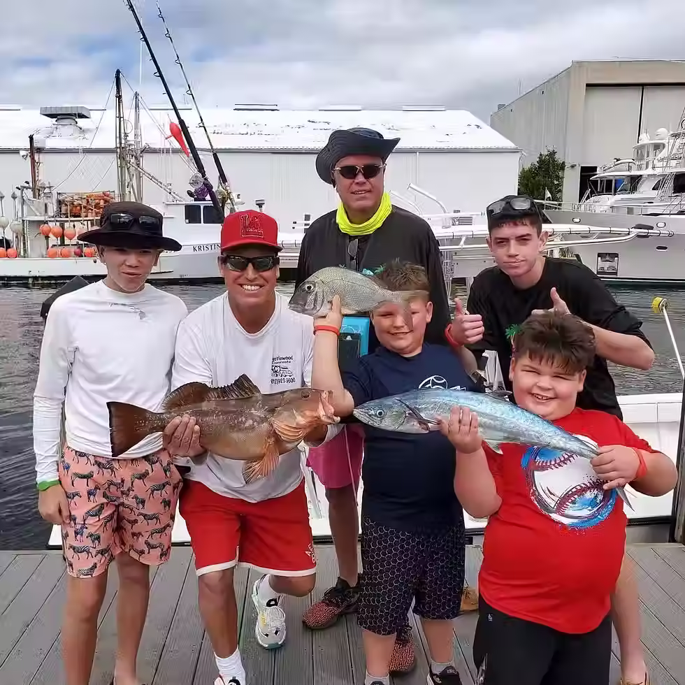 Group of six people posing with caught fish at a marina. Smiling faces and thumbs up. Boats moored in the background. Overcast sky.