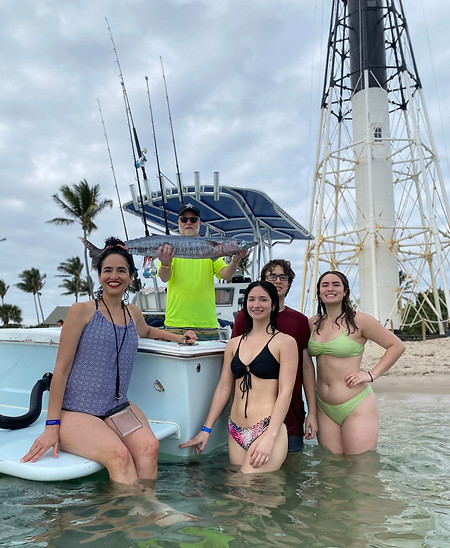 3 woman and one man standing in the wateer next to a boat while one man is standing on the boat holding up a large wahoo with a light house, palm trees, and beach in the background