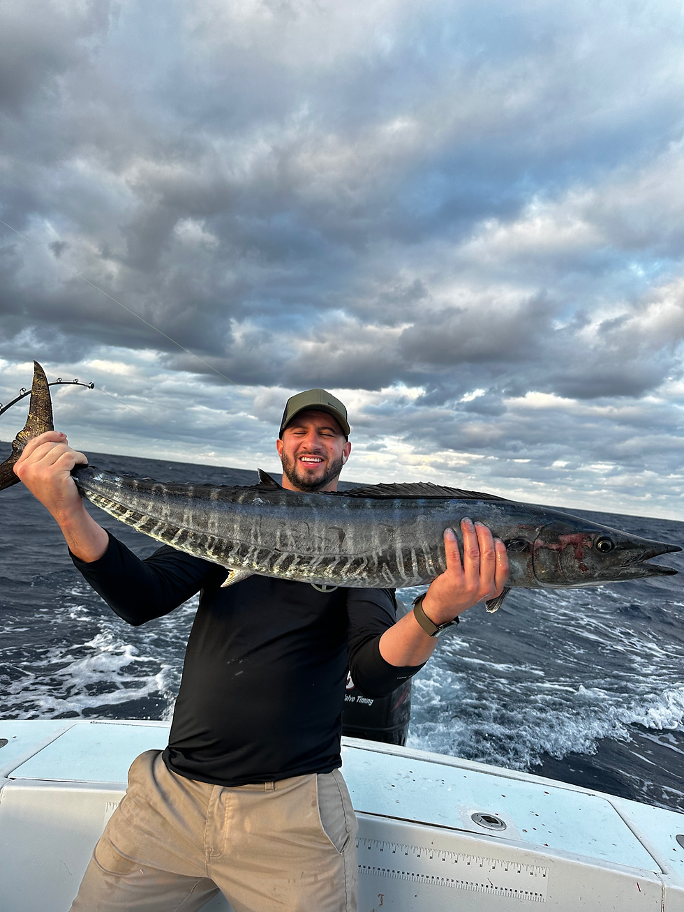 Man in black shirt and cap holds a large fish on a boat with ocean and cloudy sky in the background, smiling triumphantly.