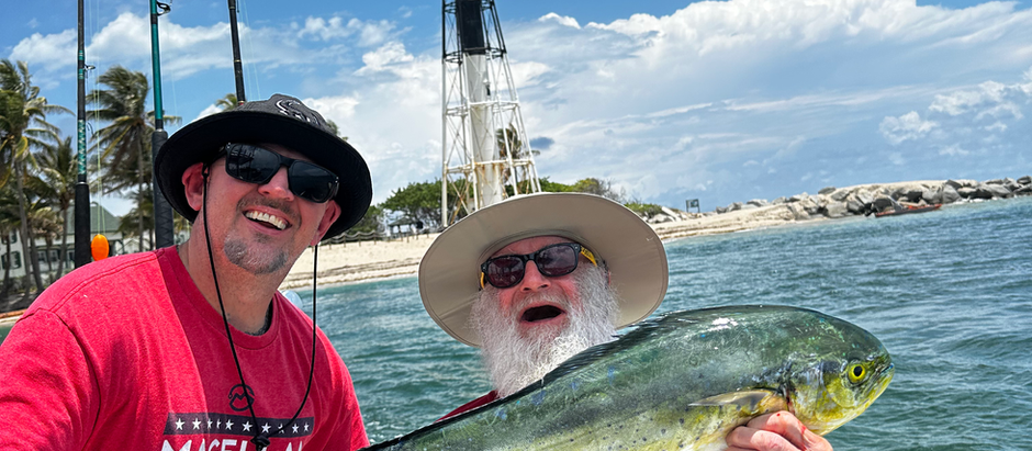 Two men on a boat hold a fish proudly. A black-and-white lighthouse stands in the background under a blue sky with white clouds.
