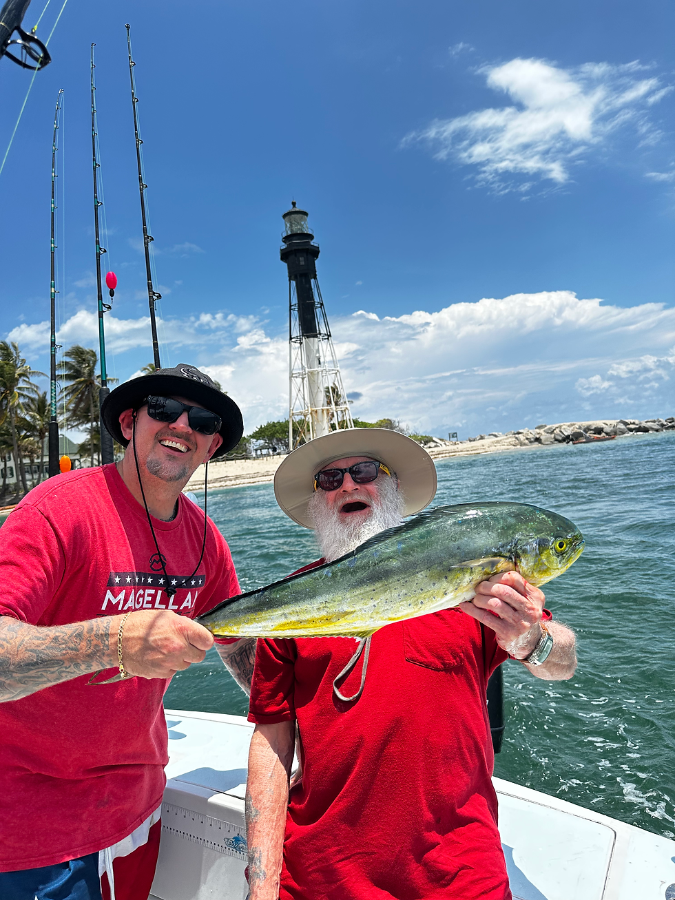 Two men on a boat hold a fish proudly. A black-and-white lighthouse stands in the background under a blue sky with white clouds.