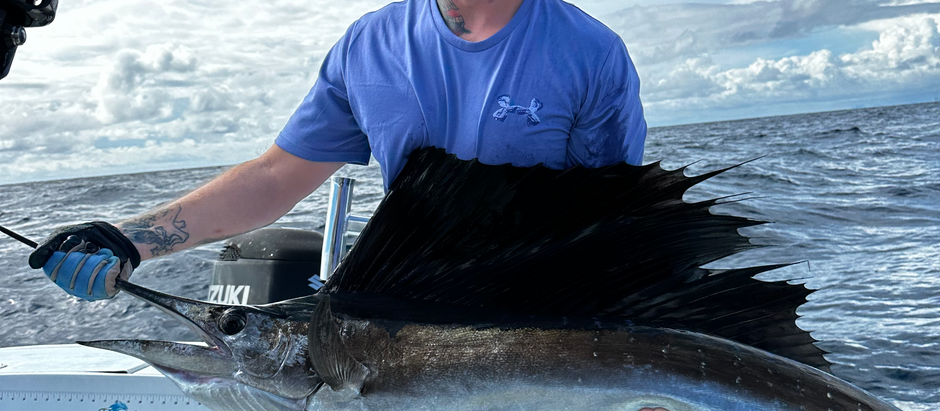 Man in sunglasses holds a large sailfish on a boat under a cloudy sky. He's smiling, wearing a blue shirt and gloves, creating a joyful mood.