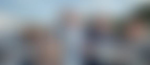 Five people on a boat hold fish, smiling under a cloudy sky. Two wear hats. Buildings and trees visible in the marina background.