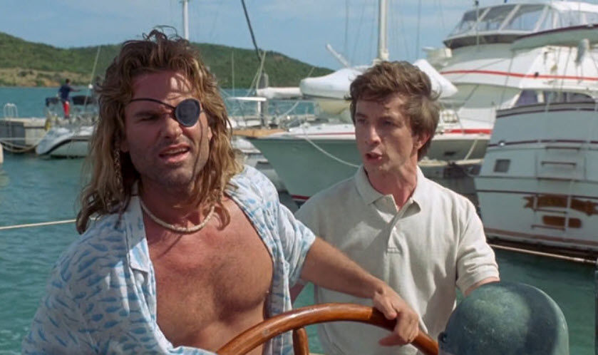Two men on a boat; one steering with an eyepatch, wearing a patterned shirt. The other, in a polo, gesturing. Yachts and hills in the background.