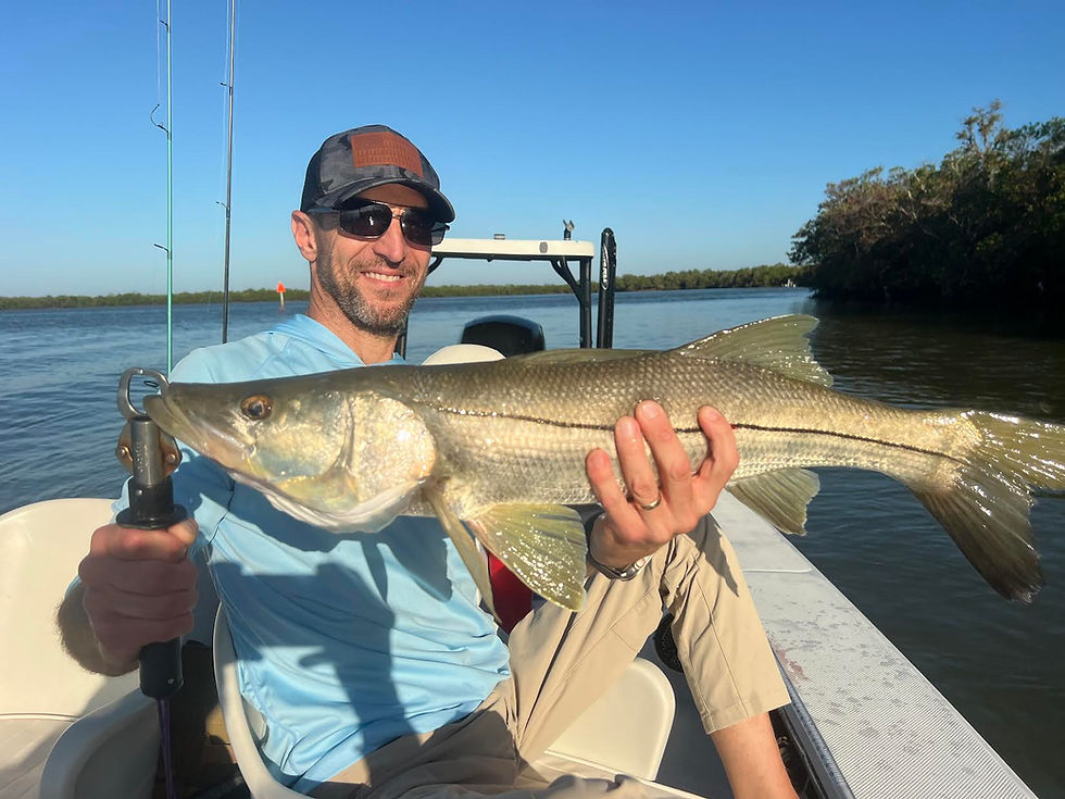 Man on a boat, smiling, holds a large fish with green and silver scales. Clear blue sky and waterway with trees in the background.