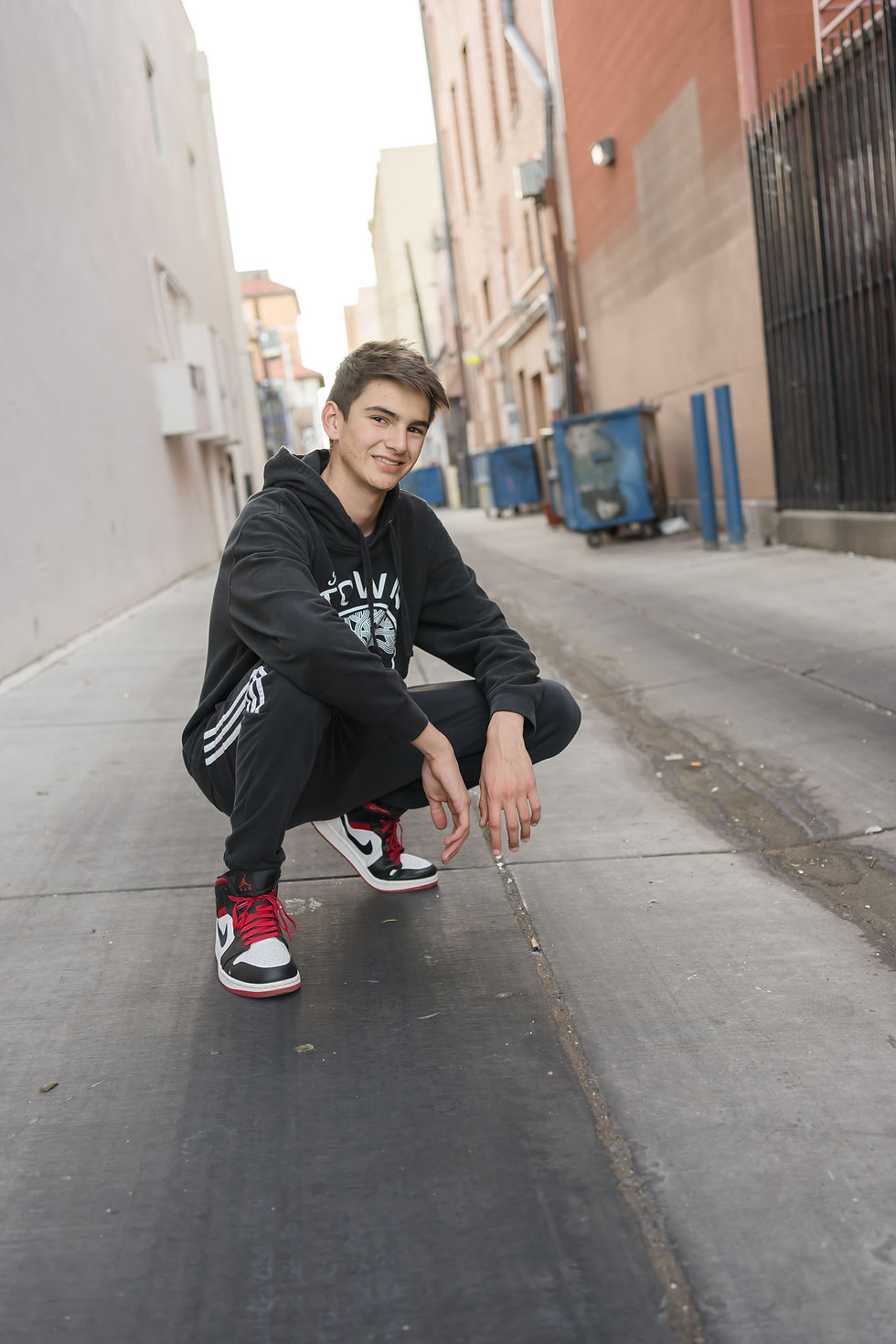 Teen squats in an alley, wearing black hoodie, striped pants, red and white sneakers. Background: urban buildings, dumpsters, relaxed mood.